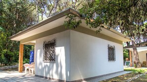 Un pequeño edificio de una sola planta con techo plano y dos ventanas con rejas metálicas. Está rodeado por un jardín con árboles y un patio empedrado con una mesa y sillas.