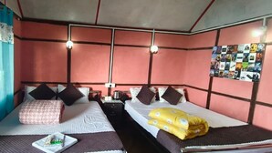 A room with two beds, each with a pillow and a blanket. There is a small table with a plate and a cup. The room has a red wall with a bulletin board and a window with curtains.