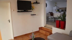 Una sala de estar con televisión, una mesa de centro y un sofá. Se ven plantas en macetas y un comedor al fondo.