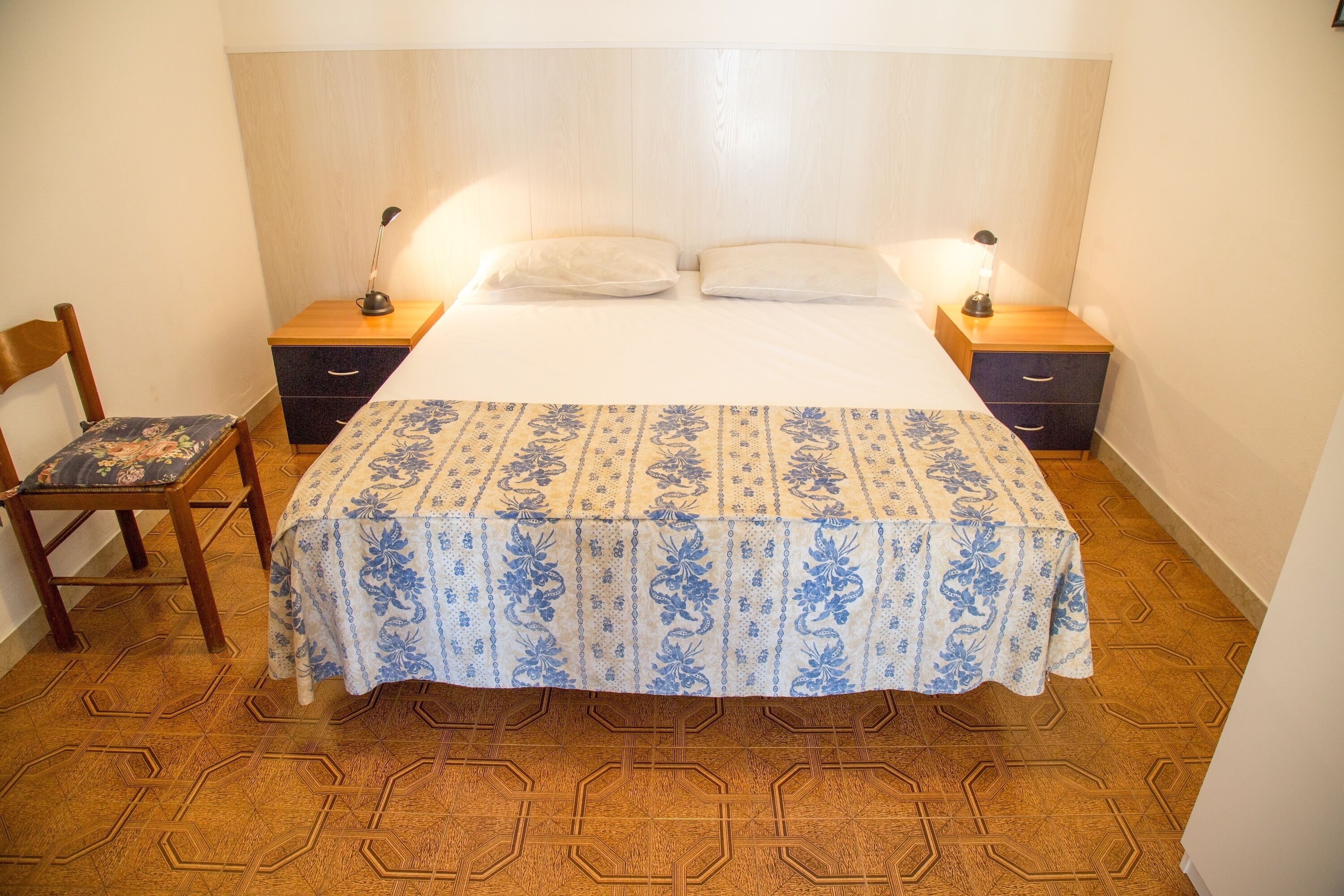 Une chambre à coucher avec une tête de lit en bois, un lit recouvert d’une couvre-lit à motifs floraux, deux tables de chevet avec des lampes, une chaise et un sol à motifs.