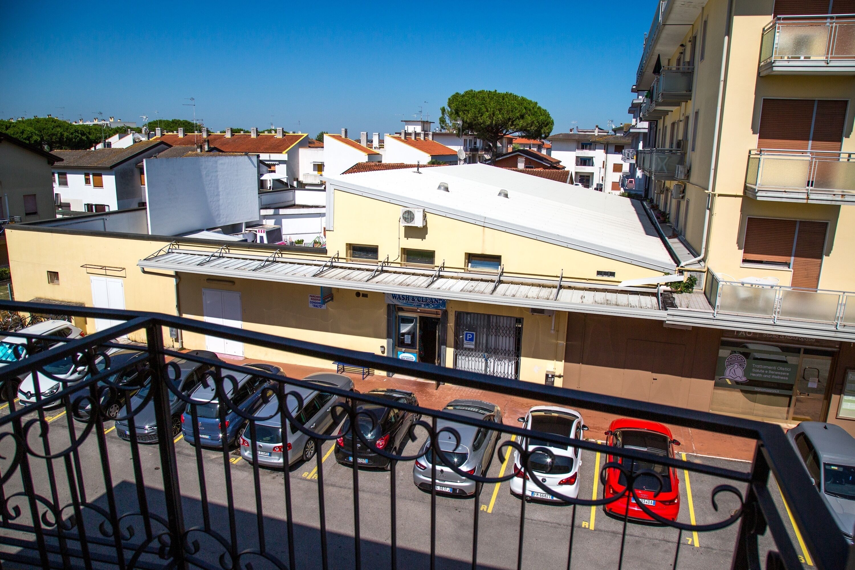 Uma área de estacionamento com vários carros, um edifício com telhado plano e uma árvore ao fundo.