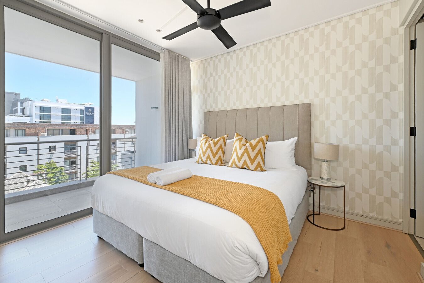 A modern hotel room with a large bed, a ceiling fan, and a view of the city through large windows.