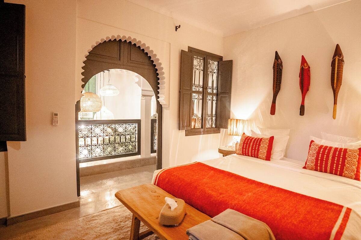 A hotel room with a bed, a bench, a table, a wall-mounted TV, a window with decorative shutters, and a door leading to an outdoor area.