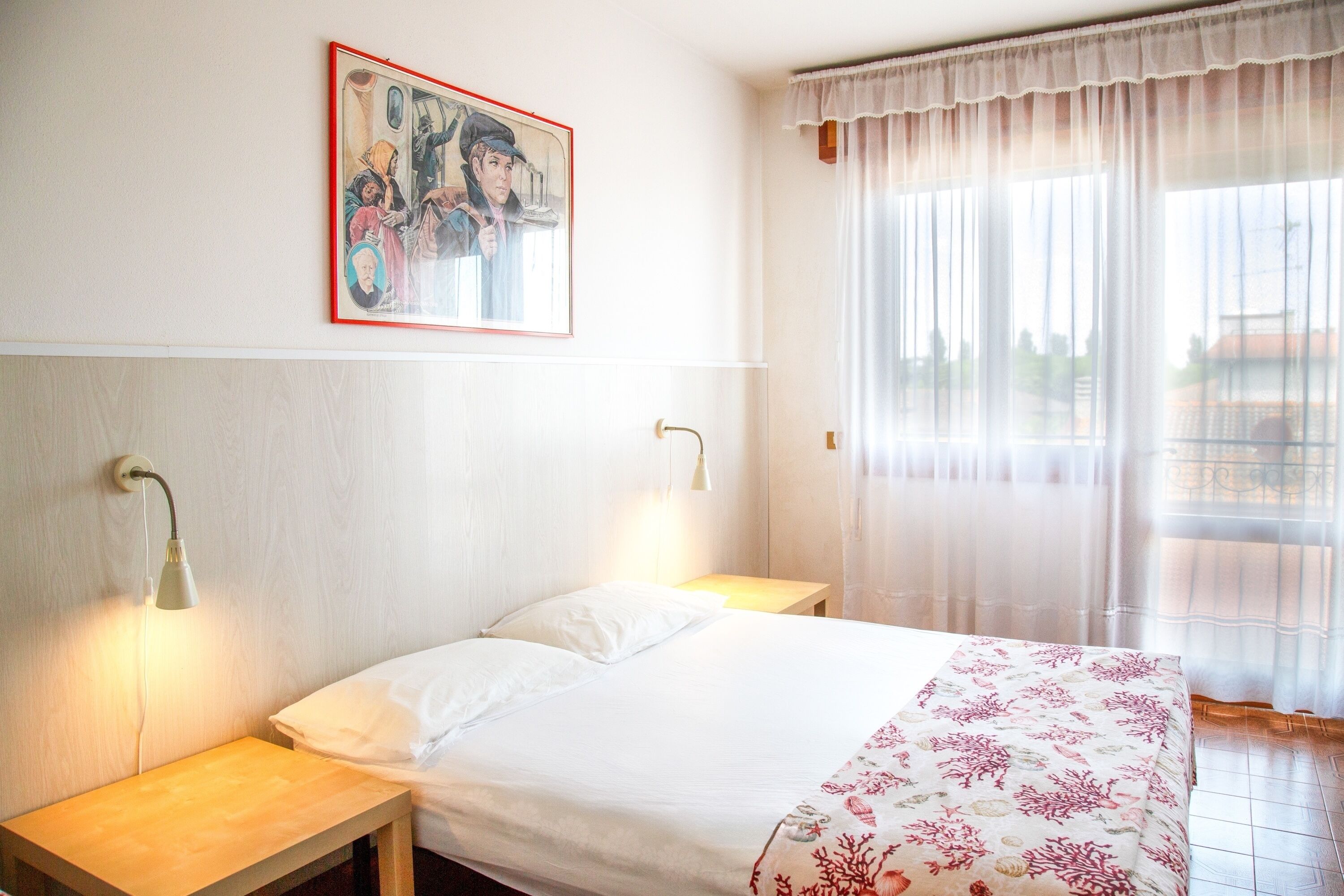 Ein Hotelzimmer mit einem Bett, zwei Nachttischen, einer Wandleuchte, einem Gemälde und einem Fenster mit durchsichtigen Vorhängen.