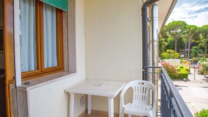 Un balcon avec une table et une chaise blanches, une fenêtre avec des volets en bois, et une vue sur une rue bordée d’arbres et dotée d’un passage piéton.