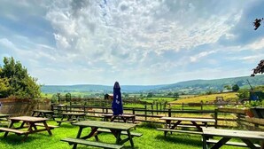 An outdoor picnic area with wooden tables and benches, surrounded by a wooden fence, overlooking a scenic landscape with hills and fields.