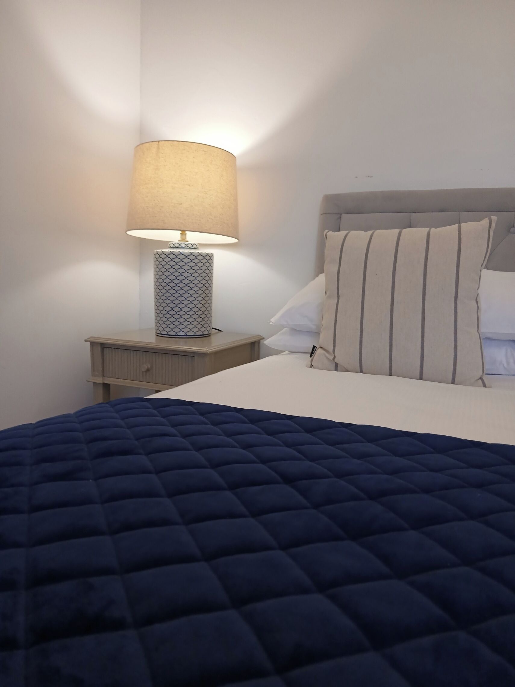 A neatly made bed with a blue quilt, a bedside lamp with a patterned shade, and a nightstand with a drawer.