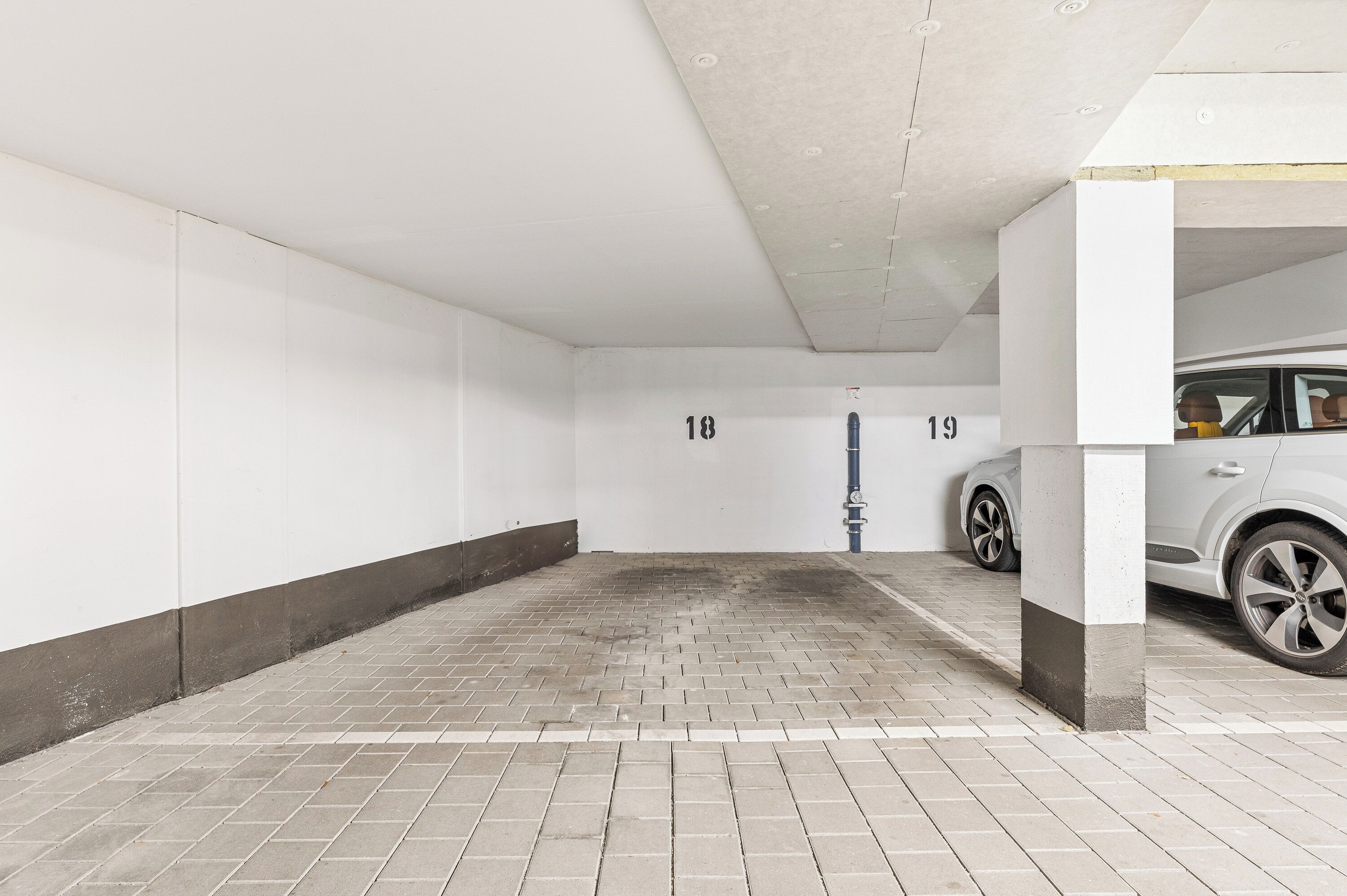 Garagem de estacionamento limpa e vazia, com vagas numeradas e um carro branco estacionado na vaga 19.