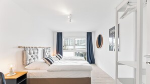 A modern bedroom with a large bed, a bedside table with a lamp, and a view of the city through a window. - Design apartment near Stuttgart/Airport (Leinfelden-Echterdingen)