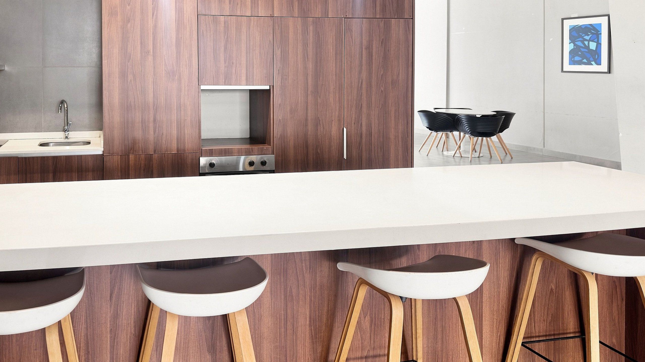 A modern kitchen with a white countertop, wooden bar stools, and a dining area with black chairs.
