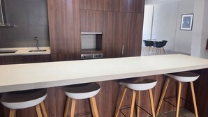 Une cuisine moderne avec un long comptoir blanc, des armoires en bois et quatre tabourets.