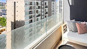 A balcony with a glass railing overlooking a cityscape, a cushioned seating area with pillows, and a textured wall. - Oceana Suites Irarrazaval (Santiago)