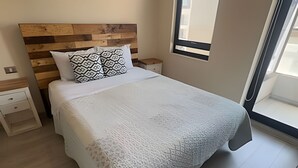 Een net opgemaakt bed met een kussen met dessin, een houten hoofdbord, een nachtkastje en een raam met jaloezieën.