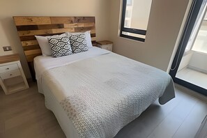 Una cama bien hecha con un cojín estampado, un cabecero de madera, una mesita de noche y una ventana con persianas.