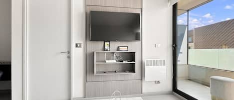 A modern living room with a flat-screen TV mounted on the wall, a small shelf with a picture frame, and a glass door leading to an outdoor balcony.