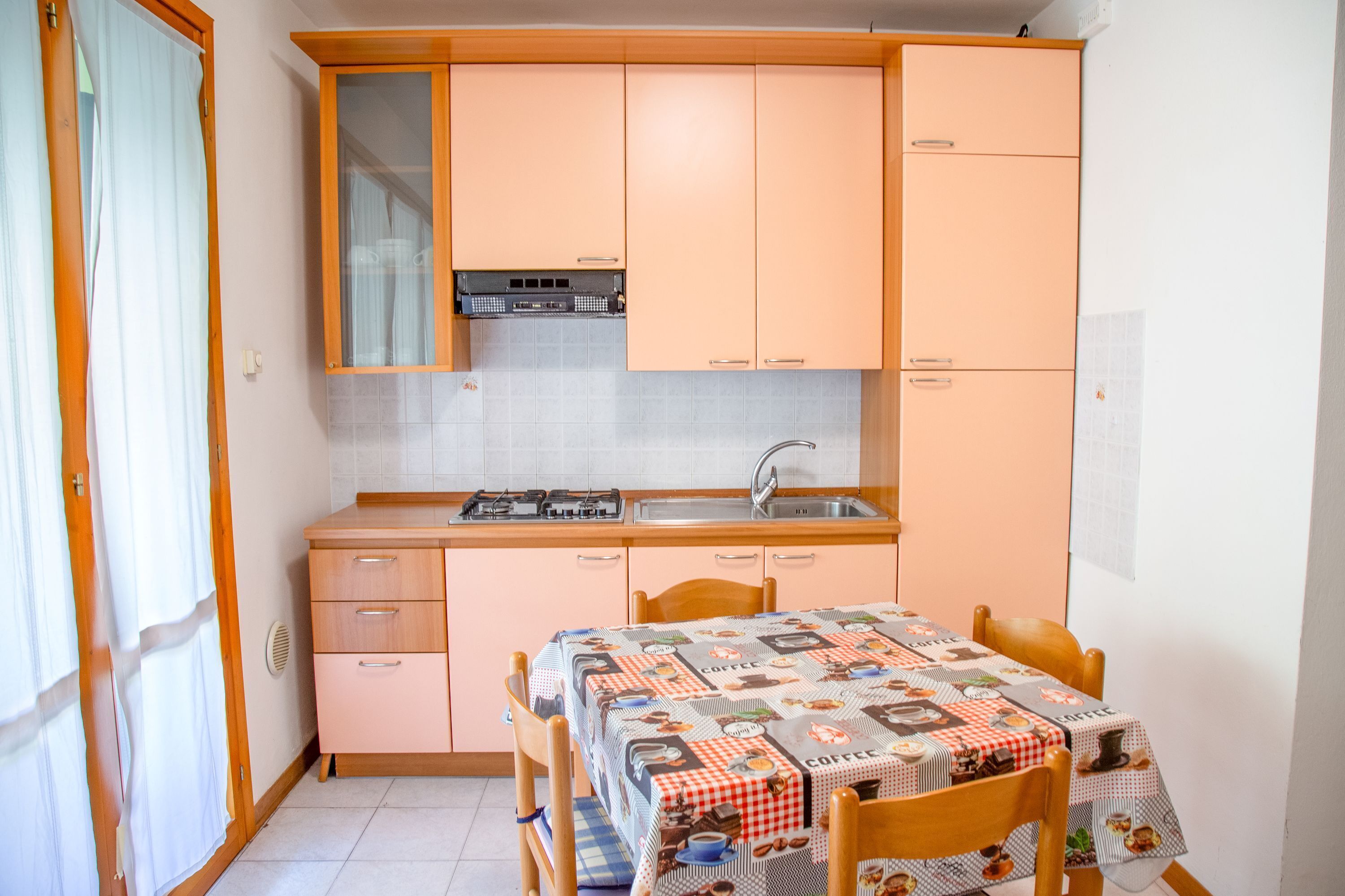A compact kitchen with wooden cabinets, a gas stove, and a sink. There is a dining table with a patterned tablecloth and two chairs.