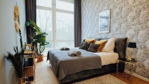 A modern bedroom with a large bed, a side table, a potted plant, and a window with curtains. - 2 room apartment in Hamburg for 6 people (Hamburg)