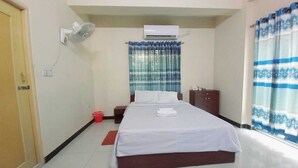 A hotel room with a bed, a bedside table, a wardrobe, a window with curtains, an air conditioner, and a wall-mounted fan. - Unique Village  (Ishurdi)