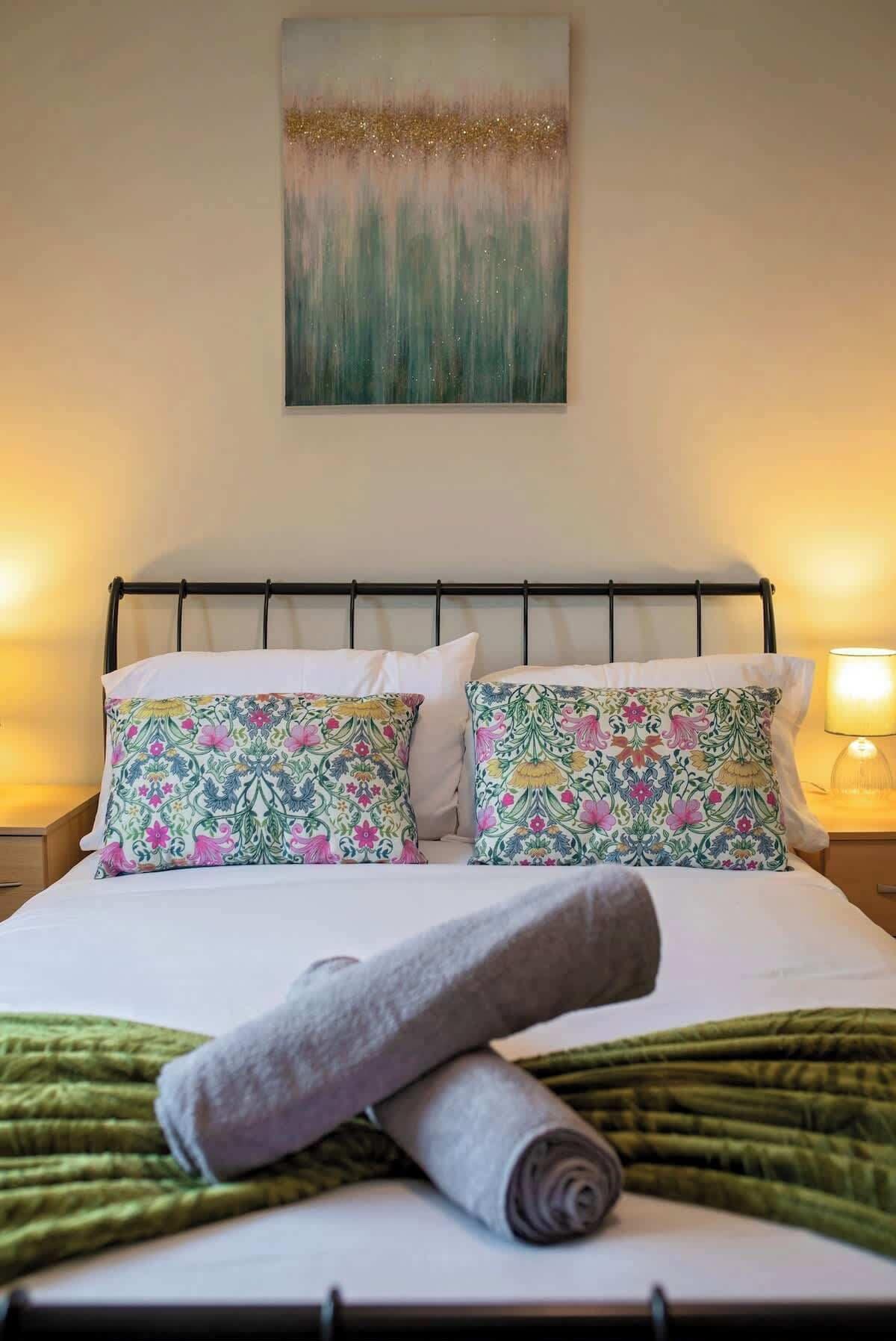 A neatly made bed with floral pillows, a green throw blanket, and a painting above.