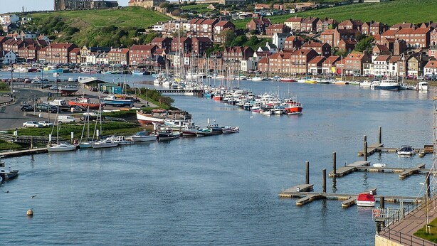 Ein Hafen mit zahlreichen vertäuten Booten, ein historisches Gebäude im Hintergrund und ein dicht bebautes Wohngebiet mit roten Dächern.