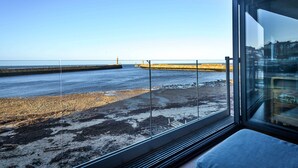 A room with a large window overlooking a beach and a pier. - No.3 Sandside (Whitby)