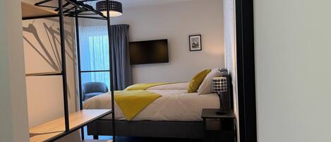 A modern hotel room with a bunk bed, a desk, a chair, a bedside table, a lamp, and a TV mounted on the wall.