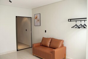 Une pièce avec un canapé en cuir marron, un porte-manteau avec des cintres, un tableau encadré au mur et une porte donnant sur une autre pièce.