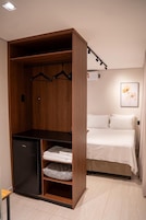Una habitación compacta con cama, armario con estantes y un refrigerador pequeño.