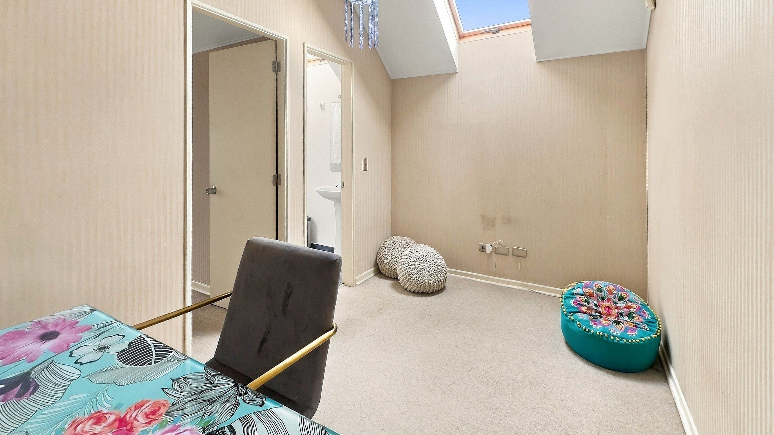 A small, narrow room with a bed, a chair, a round ottoman, and a door leading to another room.