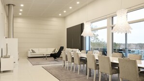 Un espace de repas moderne avec une longue table et des chaises, un canapé blanc et de grandes fenêtres donnant sur l’extérieur.