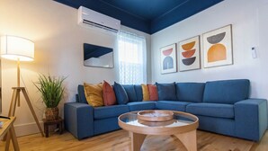 Un salon moderne avec un canapé d’angle bleu, une table basse en bois, un lampadaire, une plante en pot et des œuvres d’art abstraites accrochées au mur.