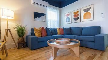 Un salon moderne avec un canapé d’angle bleu, une table basse en bois, un lampadaire, une plante en pot et des œuvres d’art abstraites accrochées au mur.