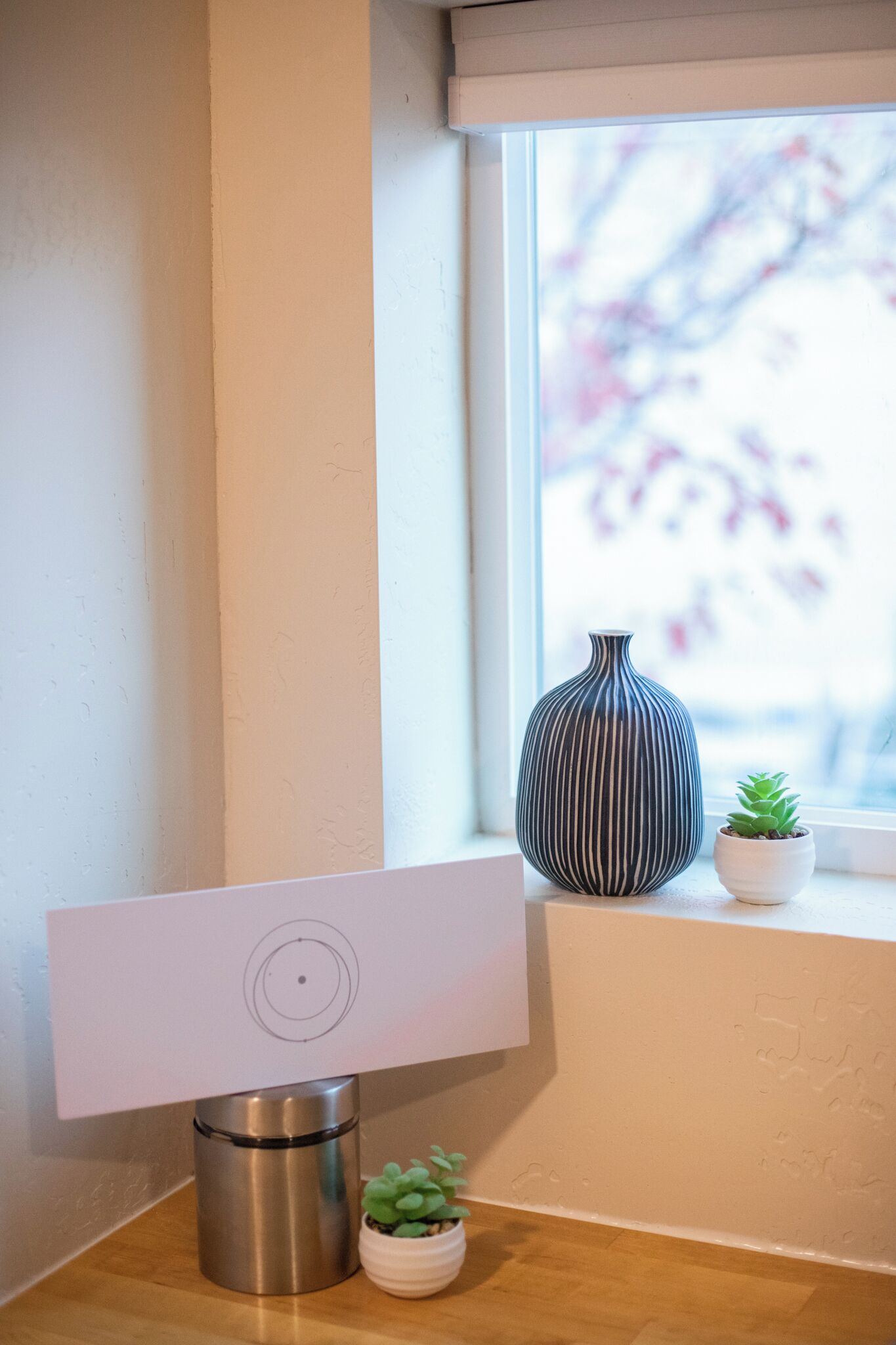 Un coin de pièce avec une lampe de bureau blanche, un objet cylindrique métallique et deux plantes en pot sur un rebord de fenêtre.