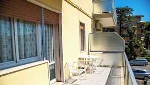 Een balkon met een tafel en stoelen, een raam met gordijnen met een patroon, en een uitzicht op geparkeerde auto's en gebouwen op de achtergrond.