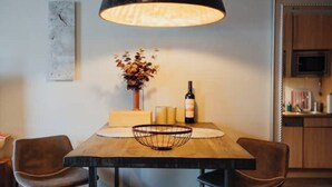 Un coin repas avec une table en bois, deux fauteuils en cuir, une suspension noire, un vase avec des fleurs séchées et une bouteille de vin.