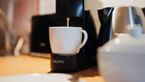 Une tasse blanche sur une machine à café Krups, une théière blanche et une assiette blanche sur une table en bois.