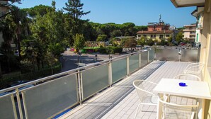 Un balcón con una mesa y sillas blancas, con vista a una calle con árboles y edificios.