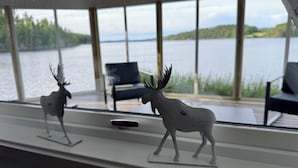 A room with a large window overlooking a lake, two moose figurines on a shelf, and a black chair.