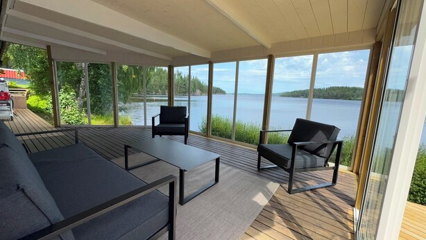 A covered deck with a sofa and chairs, large windows, and a view of a lake.