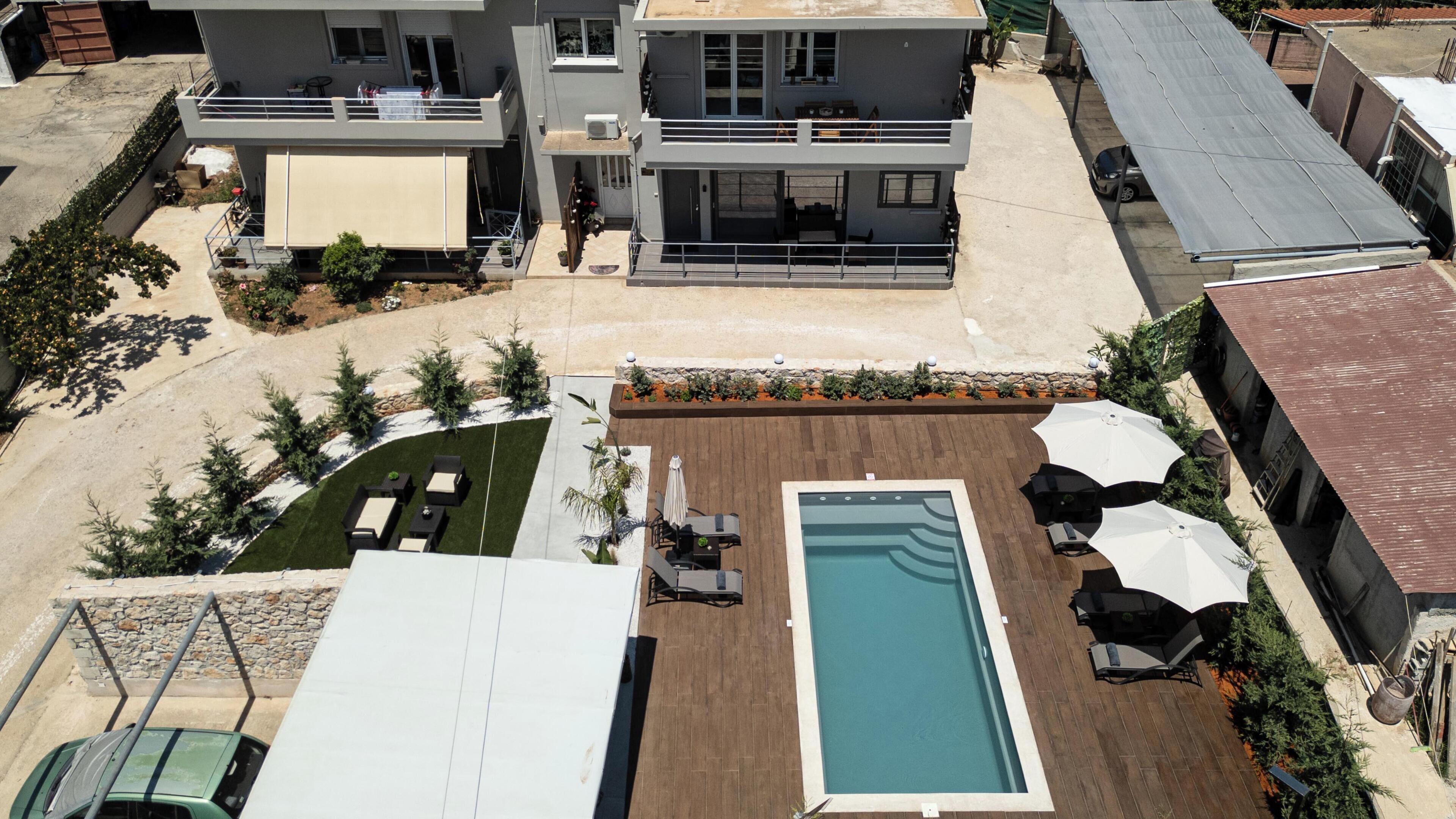 A rooftop pool area with a rectangular pool, surrounded by lounge chairs and umbrellas, and adjacent to a multi-story building with balconies.