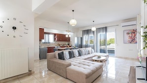 A modern living room with a beige sectional sofa, a dining table, and a kitchen in the background. - Lumina Apartment with Shared Pool (Perivolia)