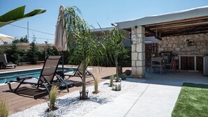 A poolside area with lounge chairs, a stone wall, and a covered outdoor kitchen. - Lumina Apartment with Shared Pool (Perivolia)