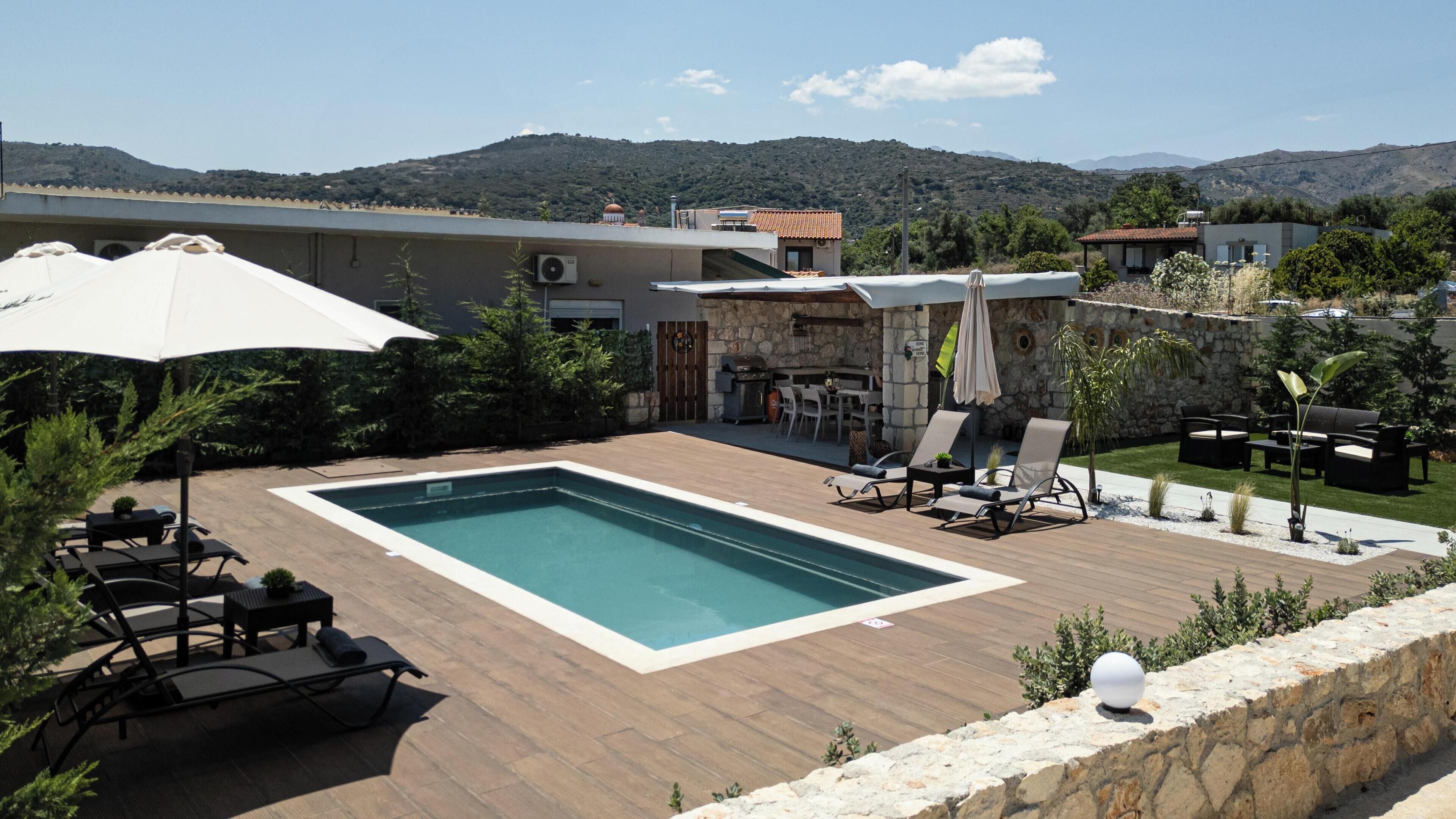 Piscina com espreguiçadeiras e guarda-sóis, cercada por um muro de pedra e vista para colinas.