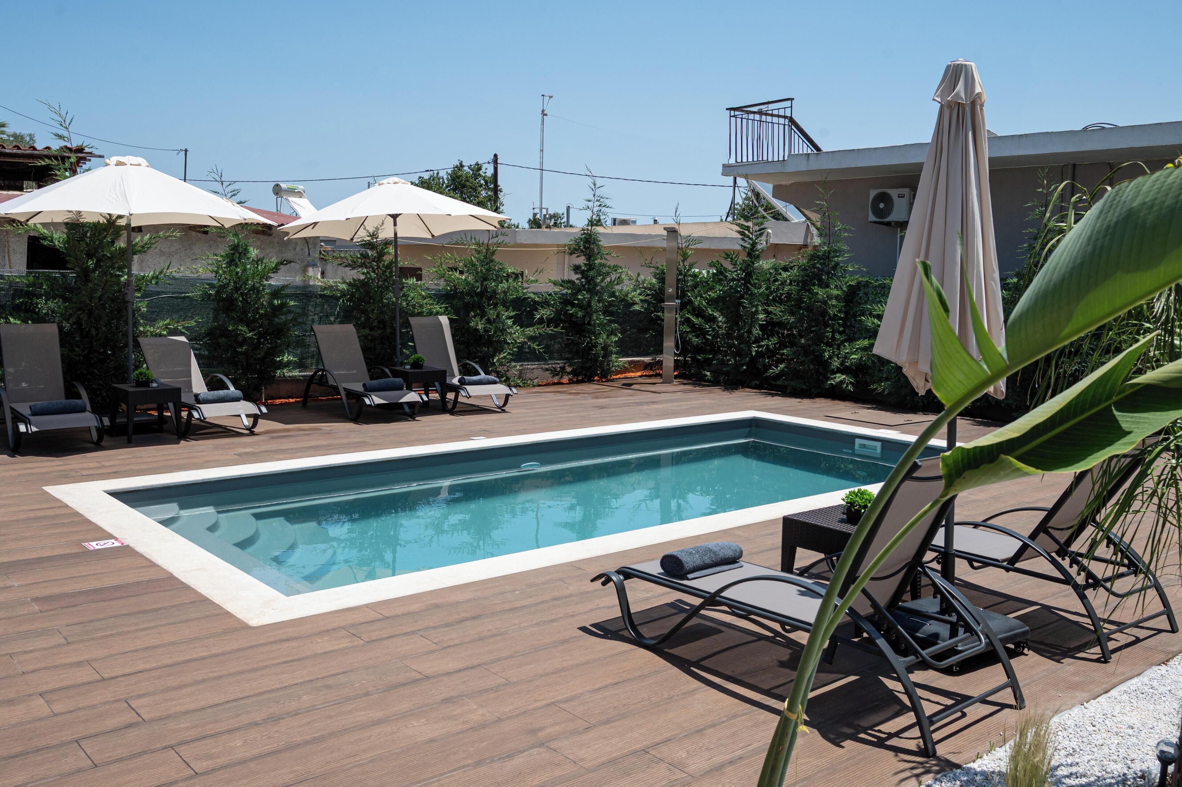 A rectangular pool with lounge chairs and umbrellas, surrounded by greenery and a building in the background.