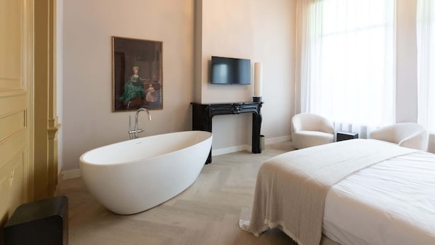 A modern hotel room with a large white bathtub, a bed with a white bedspread, a painting on the wall, a television, and a chair. - Dam Residence's Classic Suite (Zaandam)