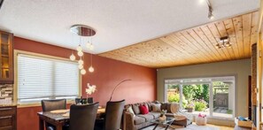 A cozy living room with a dining area, wooden ceiling, and large windows.