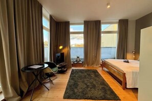 Ein Hotelzimmer mit Bett, Schreibtisch mit Stuhl, Fernseher, Fenster mit Vorhängen und Blick nach draußen.