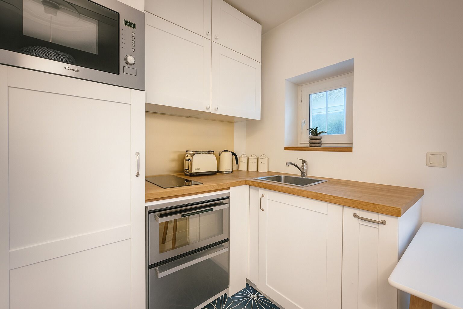 Cocina compacta con armarios blancos, microondas, tostadora, lavabo y un pequeño ventanal sobre el lavabo.