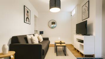 A modern living room with a dark sofa, a small wooden table, a TV on a white stand, and a mirror on the wall.