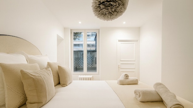 A minimalist bedroom with a large bed, a radiator, and a window with a view of buildings. - Charmant 1 Chambre à Grenelle (Paris)